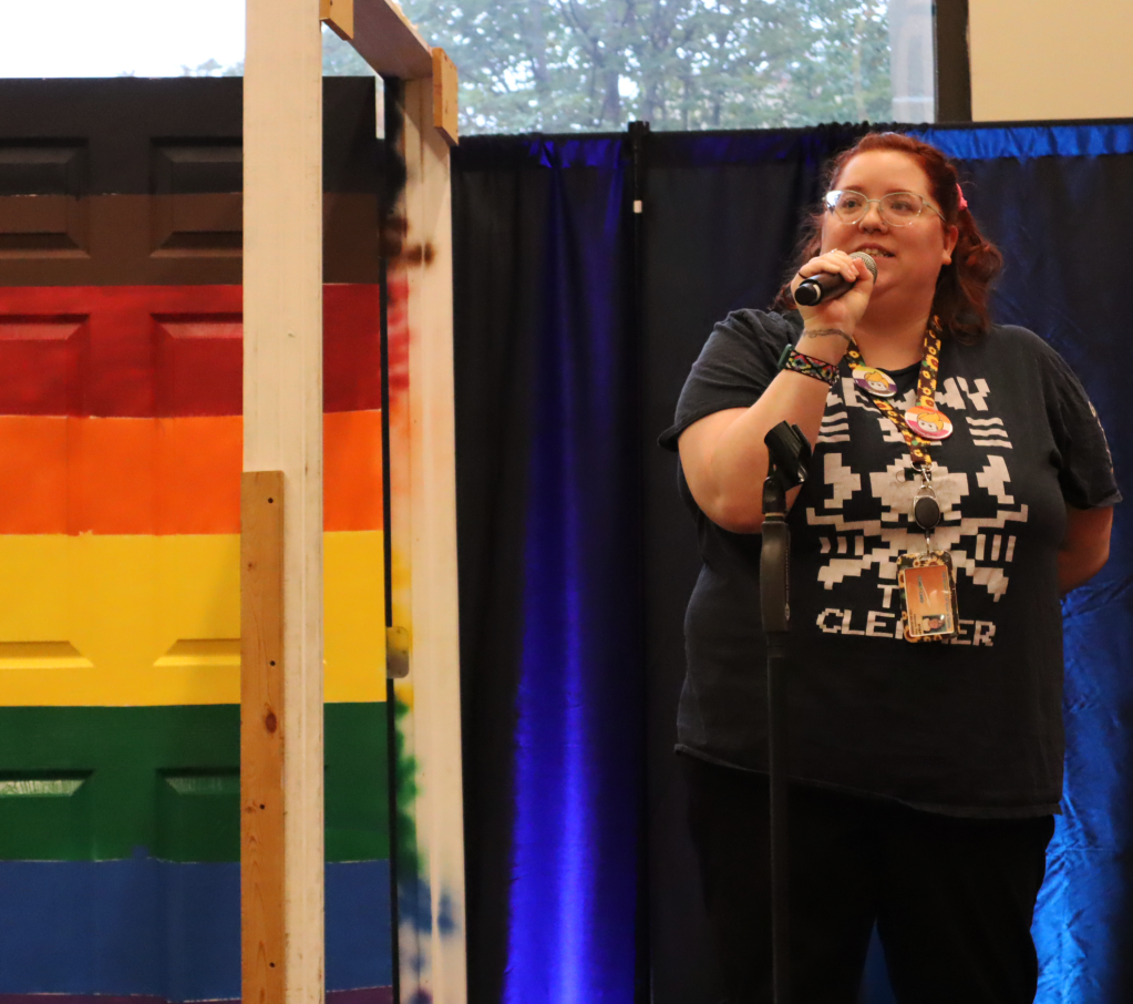 A white nonbinary person with red curly hair and blue glasses speaks into a microphone beside a free-standing door painted with the progress pride flag.